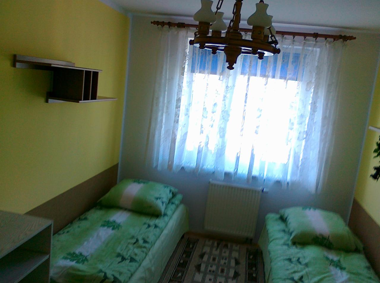 Pokój z dwoma pojedynczymi łóżkami w zielonej pościeli, drewniany żyrandol, okno z firanką i półka na ścianie. Jasne wnętrze pokoju.