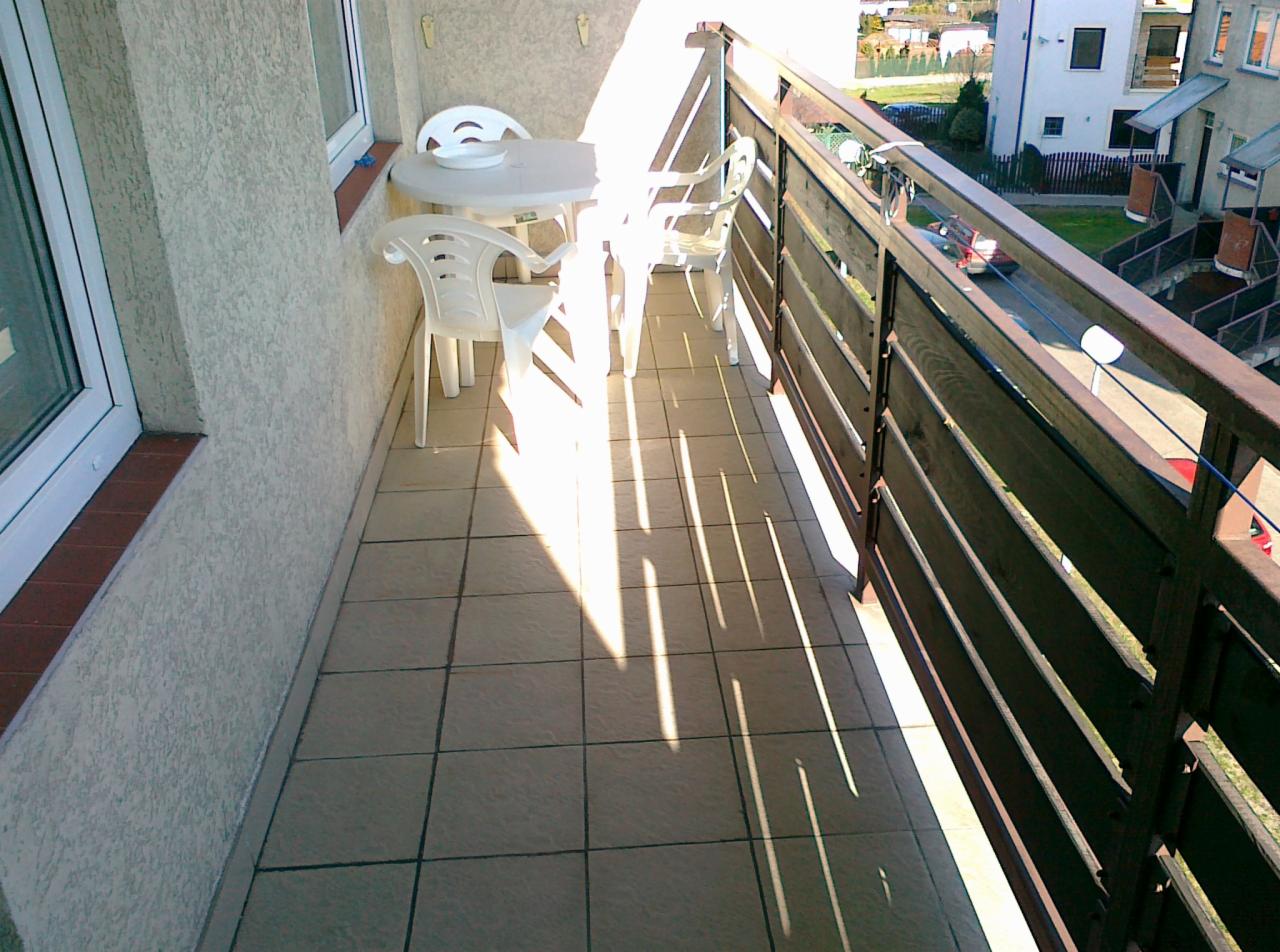 Słoneczny balkon z białymi krzesłami i stolikiem, brązowa balustrada, widok na osiedle domów. Światło słoneczne tworzy cienie na kafelkach.