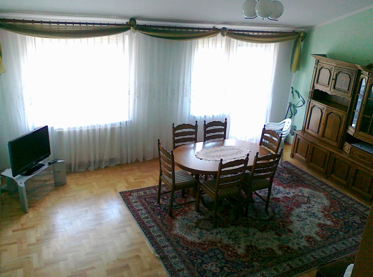 Jasny pokój z parkietem i oknami z firanami. Na dywanie okrągły stół z krzesłami, obok telewizor i kredens. W tle widać dziecięce krzesełko do karmienia.