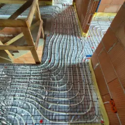 Montaż ogrzewania podłogowego w domu murowanym: białe rurki ułożone w pętle na folii izolacyjnej i siatce zbrojeniowej, widoczne fragmenty ceglanych ścian i drewnianej konstrukcji stropu.