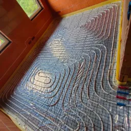 Instalacja ogrzewania podłogowego w budynku z cegły ceramicznej, rury grzewcze ułożone na siatce, widoczne okna i detale instalacyjne.