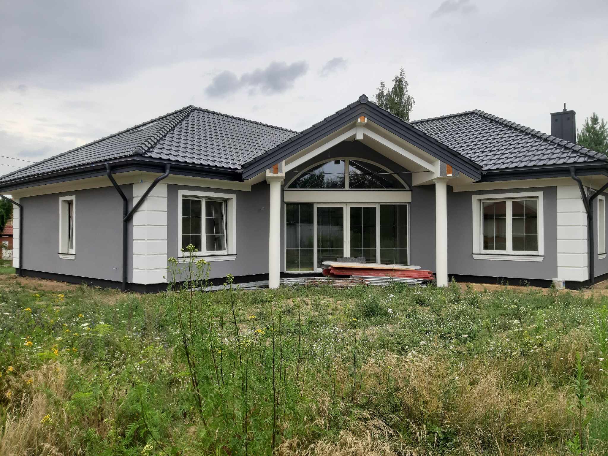 Parterowy dom z szarą elewacją, grafitową dachówką i dużym przeszkleniem w centralnej części, otoczony wysoką trawą.