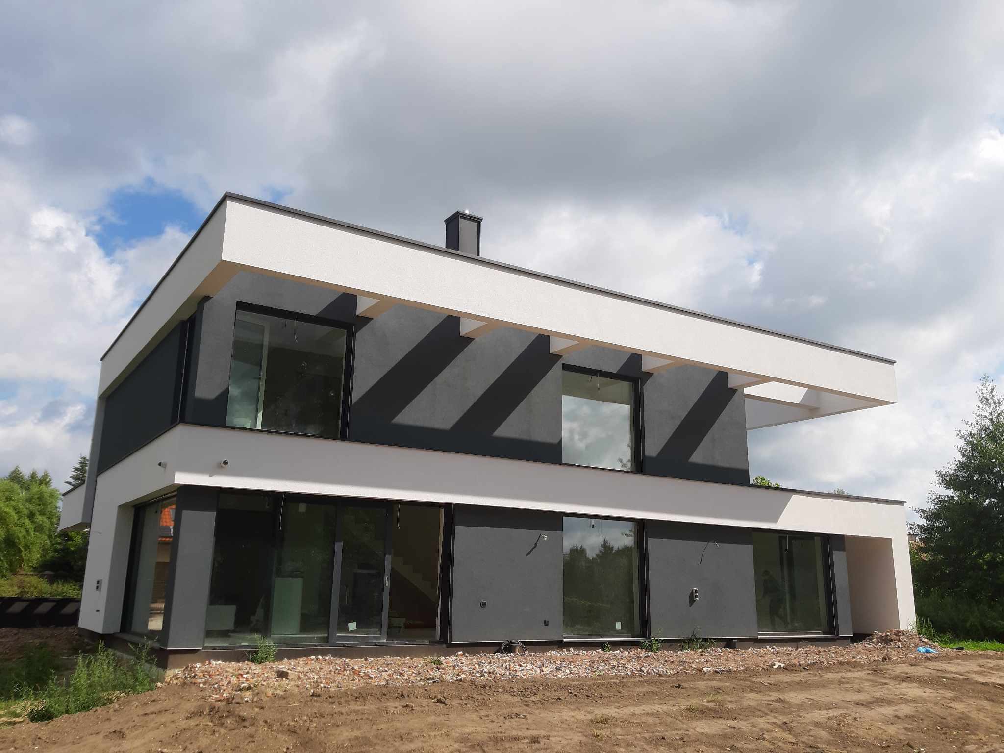 Dwukondygnacyjny dom z płaskim dachem w nowoczesnym stylu, z dużymi oknami i elewacją w odcieniach szarości i bieli, widoczny na tle pochmurnego nieba, teren wokół domu jeszcze niezagospodarowany.