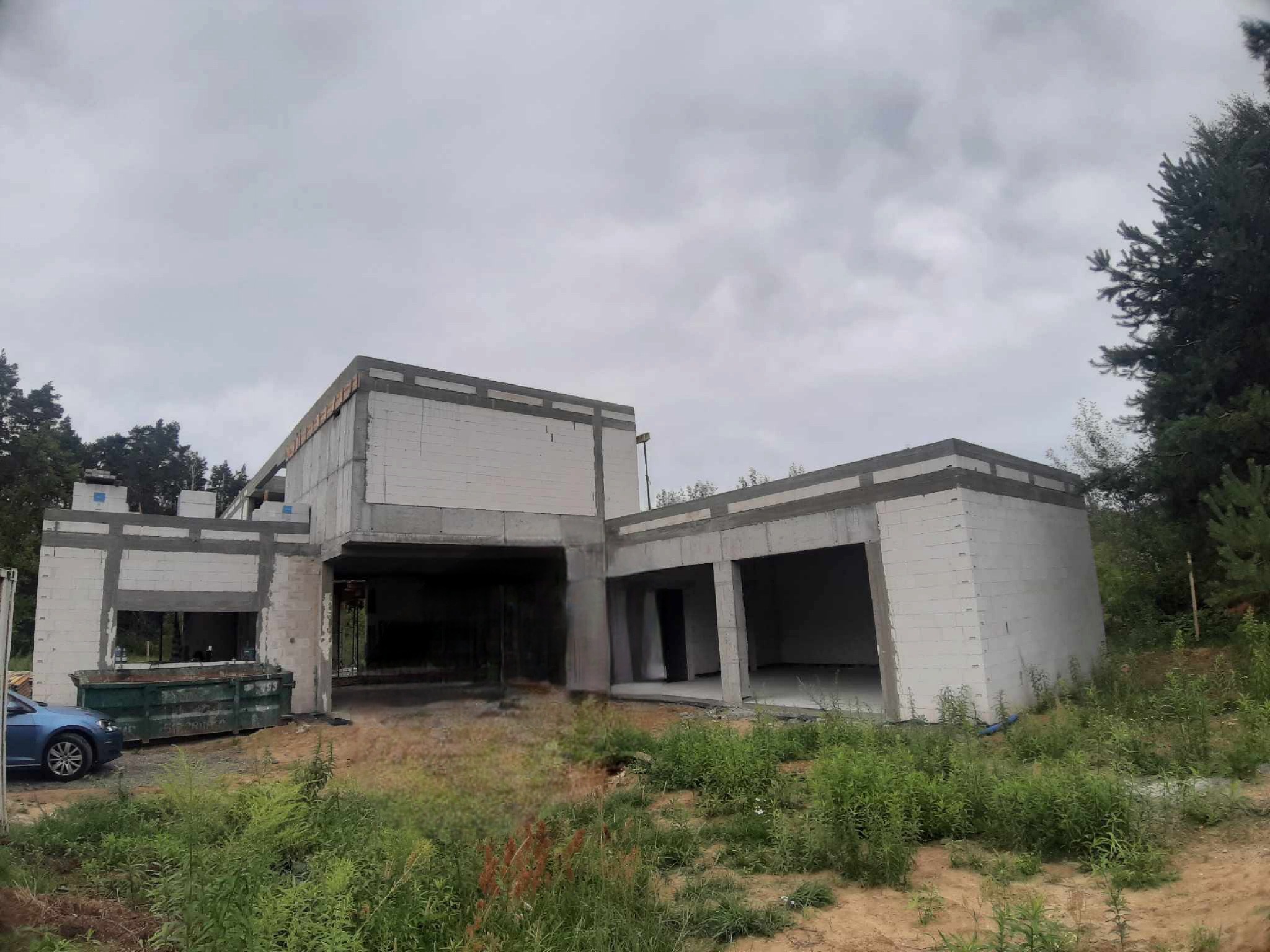 Surowy stan domu murowanego z betonu komórkowego, widoczny kontener na gruz i zaparkowany niebieski samochód osobowy.