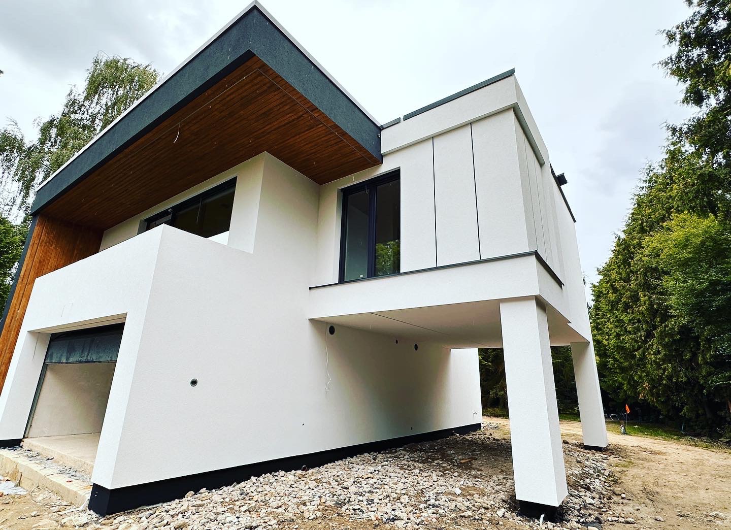 Nowoczesny, dwukondygnacyjny dom z białym tynkiem, drewnianym podbitką dachu i dużymi oknami, częściowo na podwyższeniu, widok z zewnątrz w trakcie wykończenia terenu.