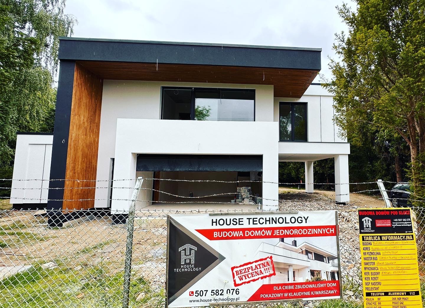 Nowoczesny, dwukondygnacyjny dom z płaskim dachem w trakcie budowy, widoczny przez ogrodzenie z siatki, z elementami drewnianej elewacji i tablicą informacyjną firmy budowlanej.