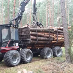 Czarno-czerwony harwester Valtra z załadowanym drewnem w lesie. Operator w kasku i kamizelce stoi obok maszyny. Widoczne wysokie sosny w tle.