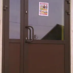 Brązowe, dwuskrzydłowe drzwi wejściowe z przeszkleniem i plakatem. Metalowa konstrukcja z uchwytem w kształcie litery D.