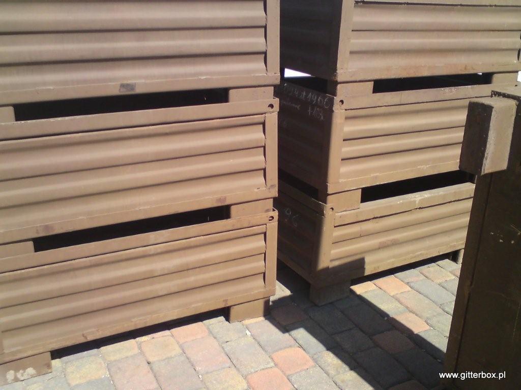 Brązowe metalowe pojemniki magazynowe ustawione jeden na drugim na kostce brukowej, z widocznymi otworami i napisami na boku.