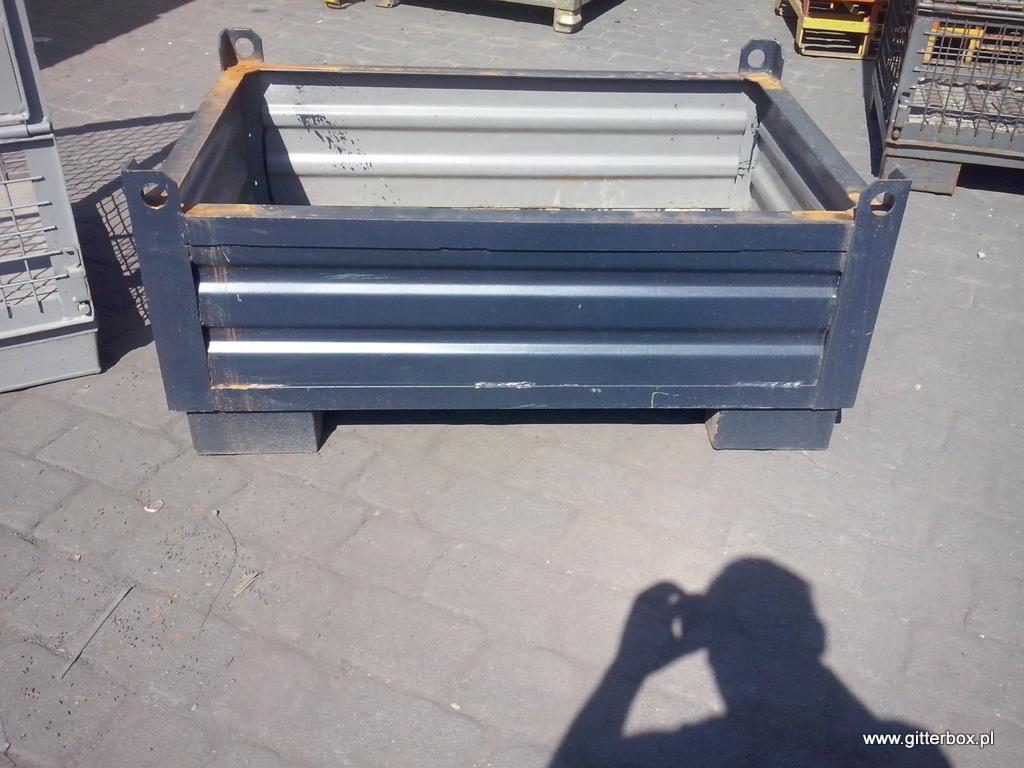 Metalowy pojemnik skrzyniowy z uchwytami, widoczny na zewnątrz w Byczynie. Solidna konstrukcja, poziome przetłoczenia, szary środek. Cień fotografa w prawym dolnym rogu.