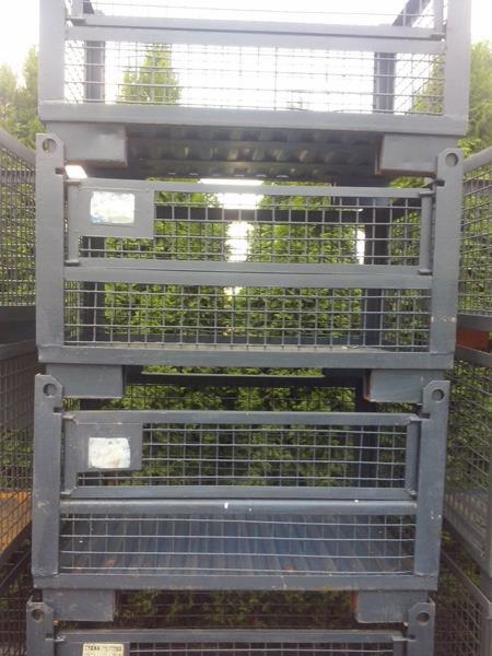 Stos metalowych, siatkowych pojemników magazynowych z szarymi ramami, ustawionych jeden na drugim na tle zieleni.