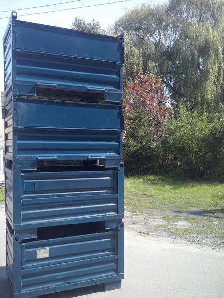 Stalowe, modułowe opakowanie transportowe w kolorze granatowym, ustawione na zewnątrz na utwardzonej powierzchni, z zielenią w tle. Widoczne otwory inspekcyjne.