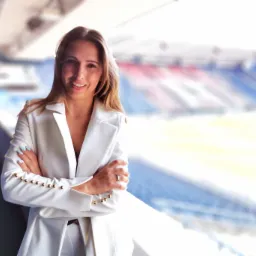 Uśmiechnięta kobieta w białym garniturze z ozdobnymi złotymi guzikami na tle rozmytego stadionu.