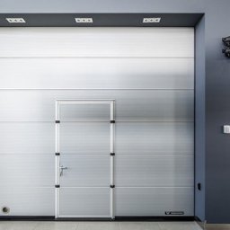 PH Podhale Sp. z o.o. - Segmentowa brama garażowa w kolorze jasnoszarym z wbudowanymi drzwiami wejściowymi, widoczna z przodu na tle szarej ściany i podłogi.