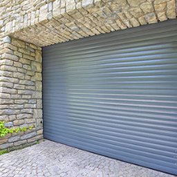 PH Podhale Sp. z o.o. - Szara brama garażowa rolowana w kamiennej elewacji, z bluszczem po prawej stronie. Kostka brukowa na podjeździe.