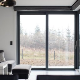 PH Podhale Sp. z o.o. - Nowoczesne, przesuwne okno tarasowe w salonie z widokiem na zewnątrz. Szara rama okienna, minimalistyczny design, jasne wnętrze. Widoczny fragment sofy i stolika.