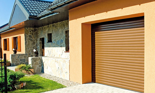 Beżowa brama rolowana garażu w budynku z elewacją z kamienia i jasnego tynku, widok z zewnątrz, w tle trawnik i fragment ogrodzenia.