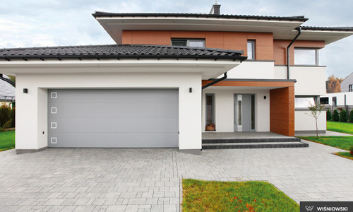 Nowoczesny dom jednorodzinny z szarą segmentową bramą garażową z czterema kwadratowymi przeszkleniami i szarymi drzwiami wejściowymi.