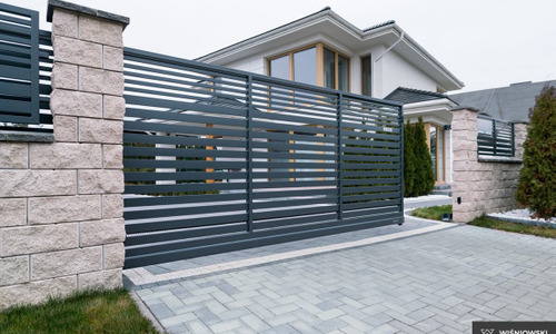 Nowoczesna, przesuwna brama wjazdowa z poziomych profili, osadzona między kamiennymi słupkami. Jasna kostka brukowa i dom w tle.