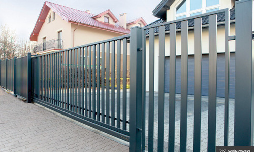 Nowoczesna, przesuwna brama wjazdowa z pionowych, metalowych profili w kolorze grafitowym, na tle domu z garażem i kostki brukowej.