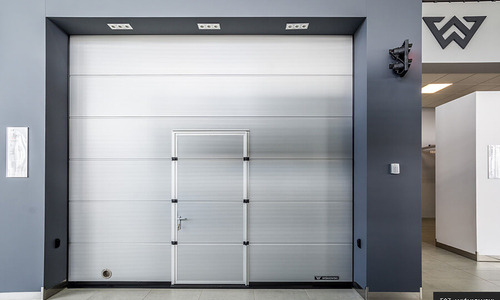 Segmentowa brama garażowa w kolorze jasnoszarym z wbudowanymi drzwiami wejściowymi, widoczna z przodu na tle szarej ściany i podłogi.