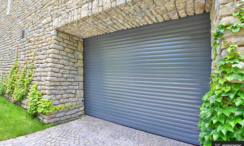 Szara brama garażowa rolowana w kamiennej elewacji, z bluszczem po prawej stronie. Kostka brukowa na podjeździe.