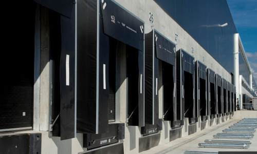 Rząd czarnych bram przemysłowych z numerami 26 i 27 na betonowej ścianie budynku magazynowego, widok z zewnątrz, w słoneczny dzień.