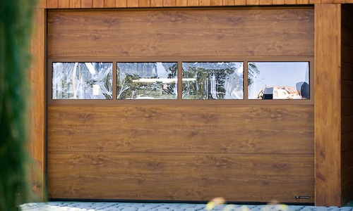 Segmentowa brama garażowa w odcieniach brązu z czterema przeszkleniami, przez które widać zieleń ogrodu i fragment budynku. Widoczne poziome panele imitujące drewno.