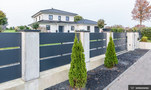 Nowoczesne ogrodzenie panelowe w kolorze antracytowym z betonowymi słupkami, na tle domu jednorodzinnego i ozdobnych tuj.