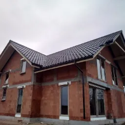 Dom w trakcie budowy z widoczną konstrukcją dachu pokrytą ciemną dachówką, ściany z czerwonej cegły ceramicznej, okna z roletami zewnętrznymi, pochmurne niebo w tle.