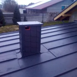 Nowy, ciemny, metalowy dach z kominem wentylacyjnym, widok z góry, w tle zabudowa jednorodzinna.