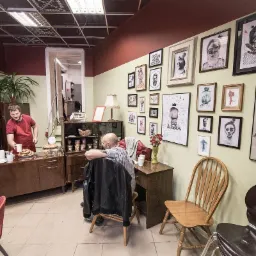 Wnętrze biura urządzone w stylu retro z meblami z lat 60., ścianą ozdobioną grafikami i plakatami, gdzie mężczyzna w czerwonej koszulce serwuje kawę i pączki, a drugi mężczyzna siedzi przy biurku.