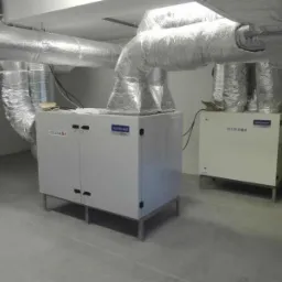 Dwa białe rekuperatory TECH-DOR z izolowanymi przewodami wentylacyjnymi w pomieszczeniu technicznym, widoczne skrzynki kartonowe w tle.