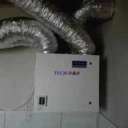 Centrala wentylacyjna TECH-DOR z podłączonymi, srebrnymi przewodami wentylacyjnymi, zamontowana na ścianie wyłożonej kafelkami.