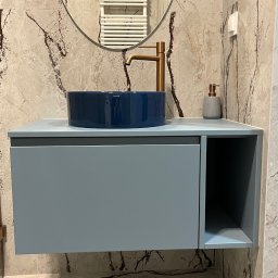 Promeb - Niebieska umywalka nablatowa SIMAS na szafce z szufladą, złota bateria, dozownik mydła na tle marmurowej ściany i okrągłego lustra z grzejnikiem.