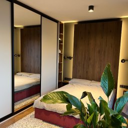 Promeb - Nowoczesna sypialnia z szafą wnękową z przesuwnymi drzwiami z lustrem, drewnianym panelem za łóżkiem i rośliną w doniczce na pierwszym planie.