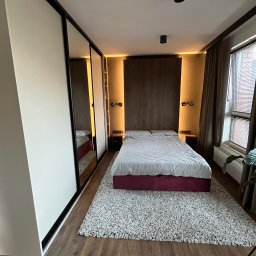 Promeb - Sypialnia z szafą przesuwną w zabudowie, drewnianym panelem za łóżkiem i dywanem shaggy. Ciemne ramy szafy i oświetlenie punktowe.