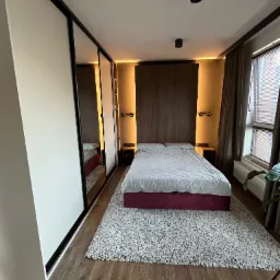 Sypialnia z szafą przesuwną w zabudowie, drewnianym panelem za łóżkiem i dywanem shaggy. Ciemne ramy szafy i oświetlenie punktowe.