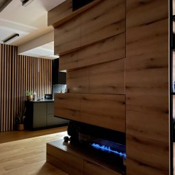 Promeb - Nowoczesna zabudowa drewniana z biokominkiem i szafkami, widok na czarną kuchnię z drewnianymi lamelami. Oświetlenie punktowe.