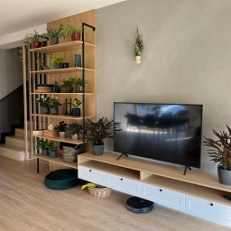 Promeb - Nowoczesny salon z drewnianą zabudową TV i regałem na rośliny, robot sprzątający. Jasne drewno, szare ściany i podłoga tworzą spójną całość. Minimalistyczny design.