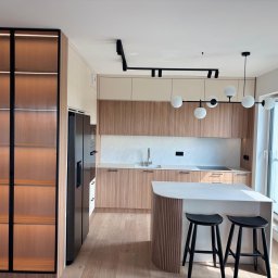 Promeb - Nowoczesna kuchnia z drewnianymi frontami, wyspą i szklaną witryną z podświetleniem LED. Minimalistyczny design, czarne akcenty oświetlenia i stołków barowych.