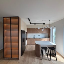 Promeb - Nowoczesna kuchnia z drewnianymi frontami, wyspą z białym blatem i hokerami. Na ścianie szafki i oświetlenie. Charakterystyczna witryna z ryflowanego szkła.