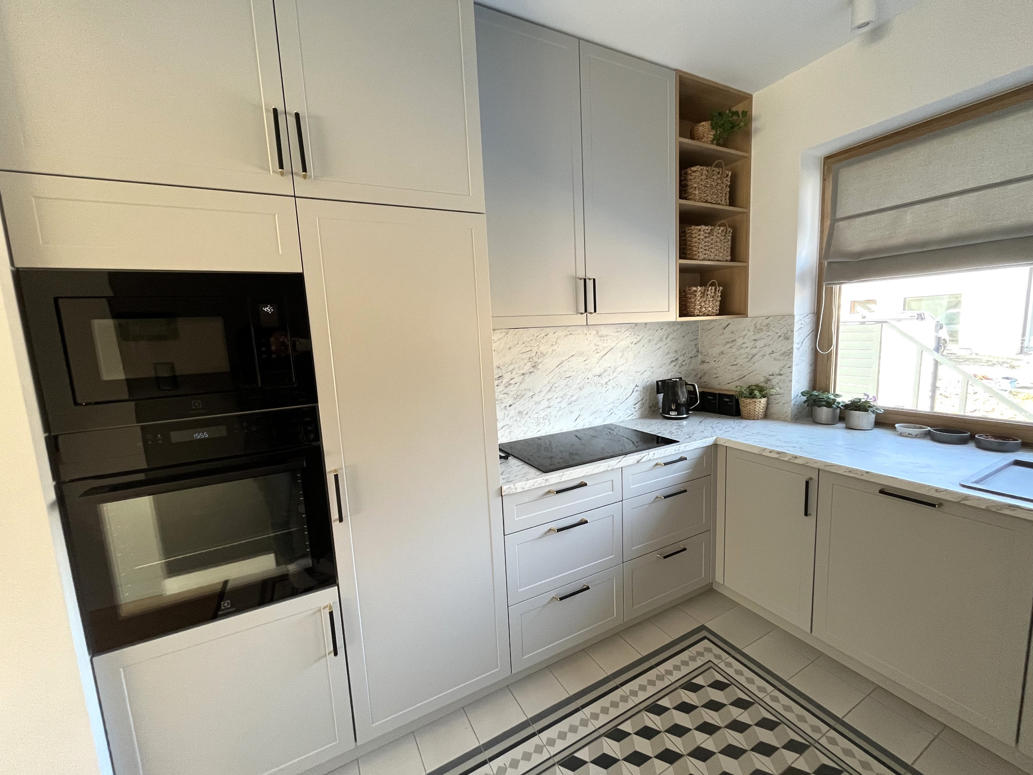 Jasnoszara kuchnia na wymiar z marmurowym blatem, czarnym piekarnikiem i płytą indukcyjną, uzupełniona drewnianym regałem z koszykami i oknem z roletą rzymską.