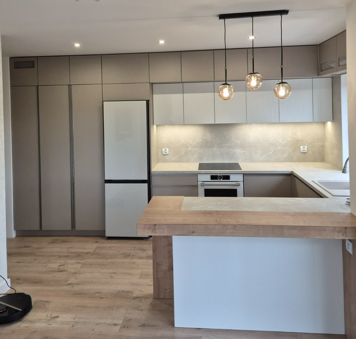 Nowoczesna kuchnia w neutralnych barwach z wyspą, oświetlona wiszącymi lampami. Fronty szafek w odcieniach beżu i bieli, drewniany blat. Widoczny sprzęt AGD.