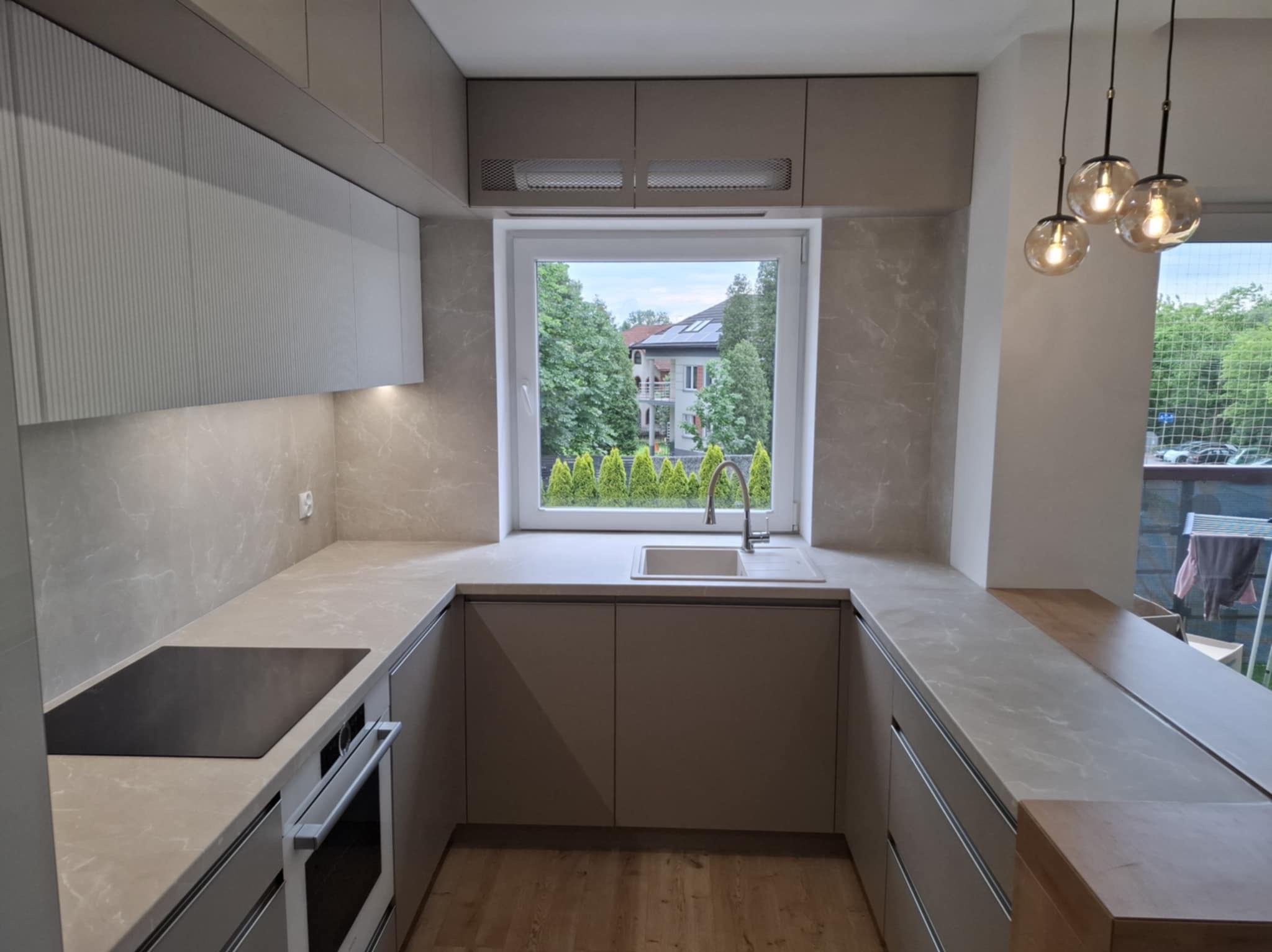 Nowoczesna kuchnia w kształcie litery U z blatem imitującym marmur, oświetlona wiszącymi lampami. Za oknem widok na zieleń i domy.