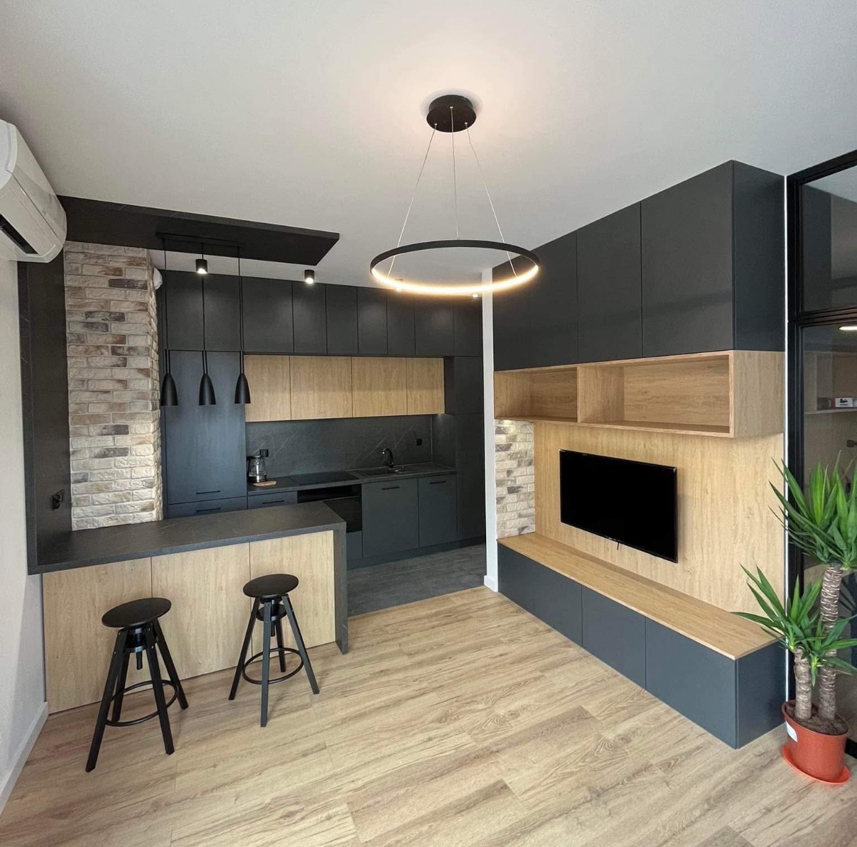 Nowoczesna kuchnia z szarymi szafkami i drewnianymi blatami, oświetlona okrągłą lampą. Dwa czarne hokery przy wyspie. Fragment ściany z cegły.