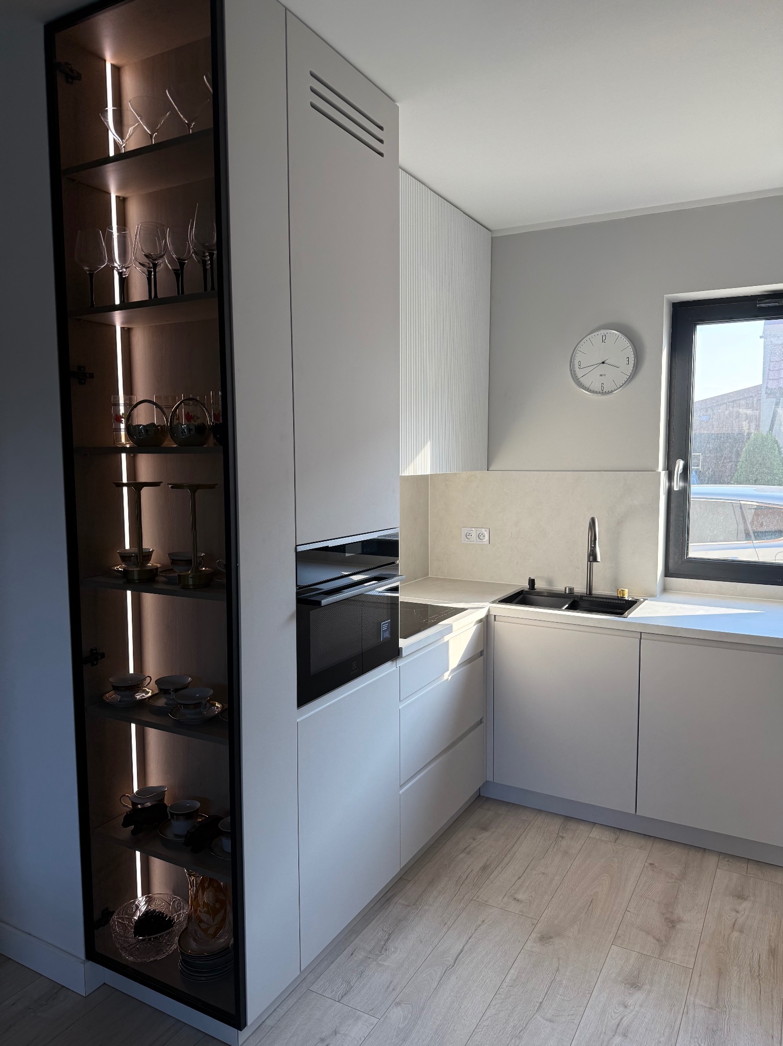 Nowoczesna, jasnoszara kuchnia z oświetloną szafką ekspozycyjną na zastawę stołową i szklanki. Minimalistyczny design, widok na zewnątrz przez okno.
