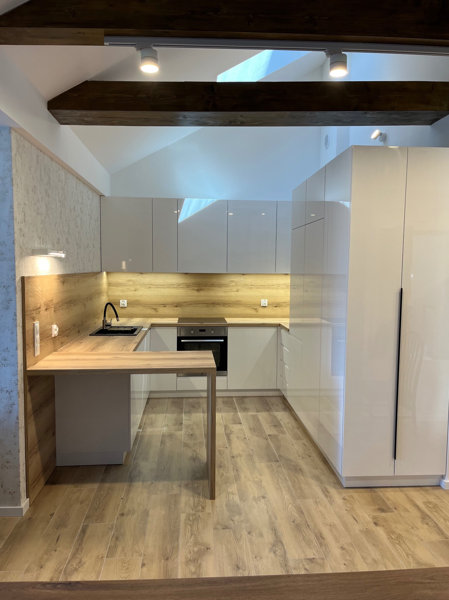 Biała kuchnia z drewnianym blatem i wykończeniem, oświetlona punktowo, widok z perspektywy podłogi. W tle drewniane belki stropowe.