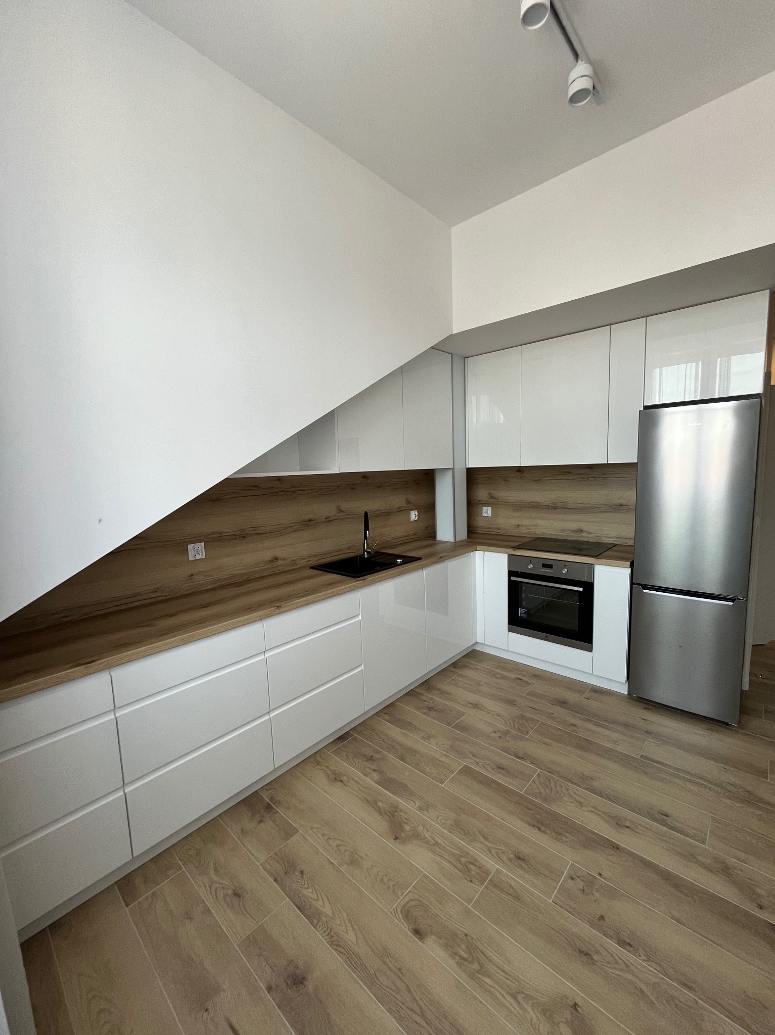 Nowoczesna kuchnia z białymi, lakierowanymi frontami i drewnianym blatem. Uwagę zwraca czarny zlew i minimalistyczne oświetlenie sufitowe.