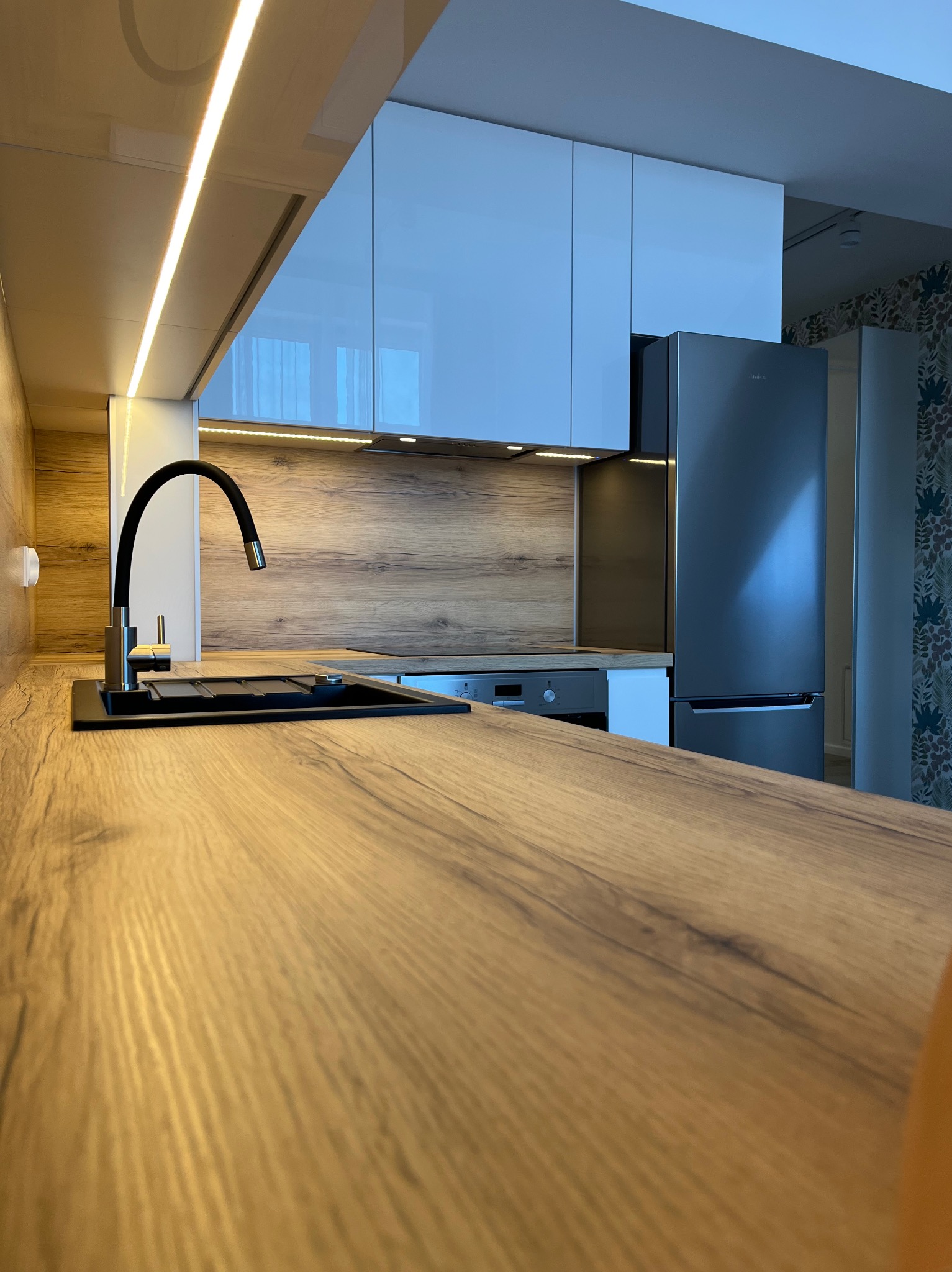 Nowoczesna kuchnia z drewnianym blatem, czarnym zlewem i baterią. Białe szafki górne z oświetleniem LED. W tle lodówka i ściana w kwiatowy wzór.